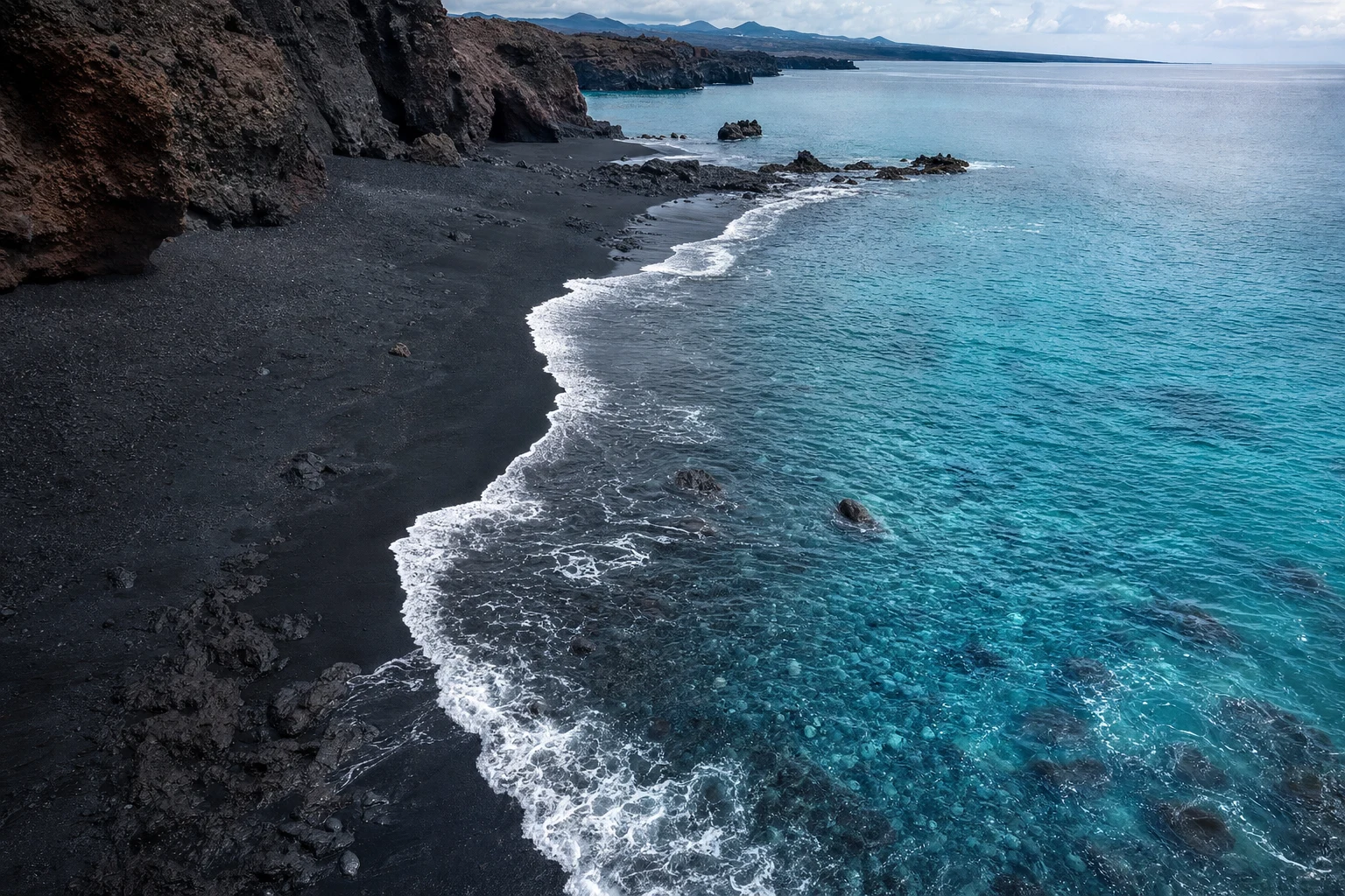 Vista aérea de una playa volcánica negra que se encuentra con agua color aguamarina clara, con detalle natural de costa y un horizonte amplio.