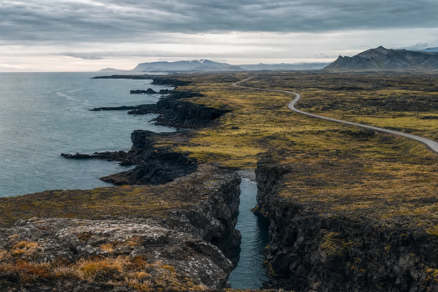 Una costa de basalto con rocas oscuras, canales de marea, una carretera interior estrecha y luz gris suave abriéndose hacia el horizonte.