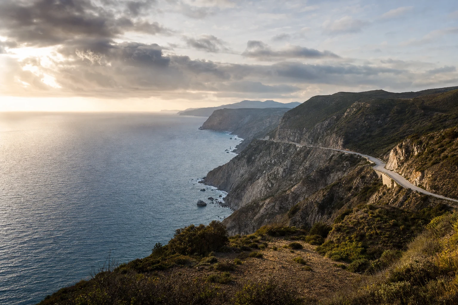 Amplio mirador costero al atardecer con acantilados en capas, un camino curvo sobre el promontorio y nubes suaves sobre un mar tranquilo bajo luz natural.
