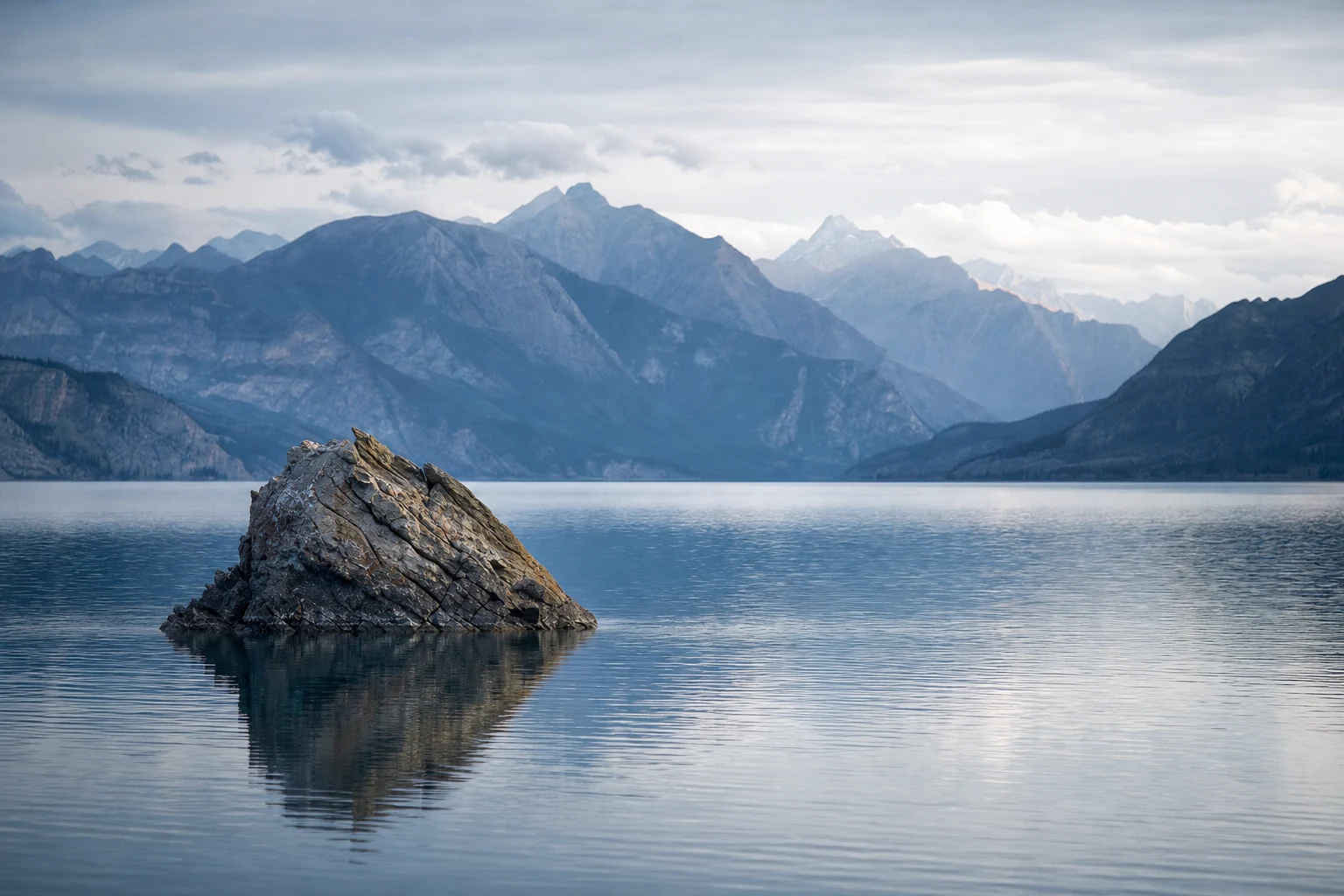 A solitary rock rising from a smooth blue lake with layered mountains in the background under soft natural light.