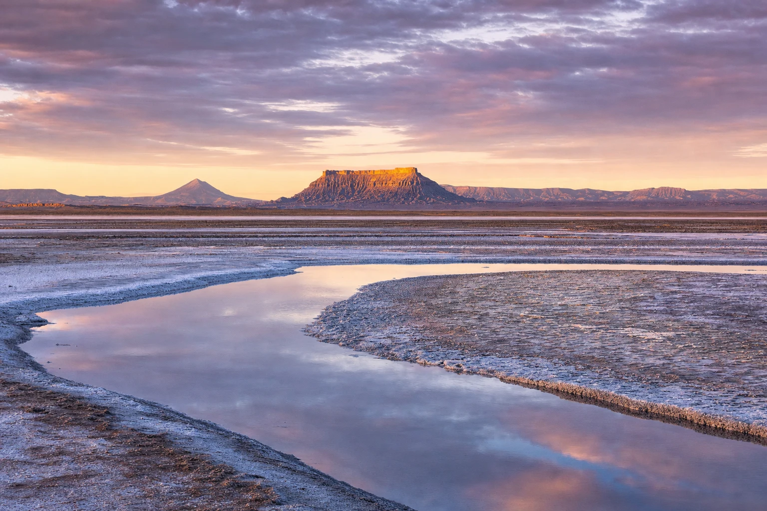 Wide salt flat at dawn with a thin reflective stream, distant mesas, soft lavender clouds, and long shadows across pale ground.