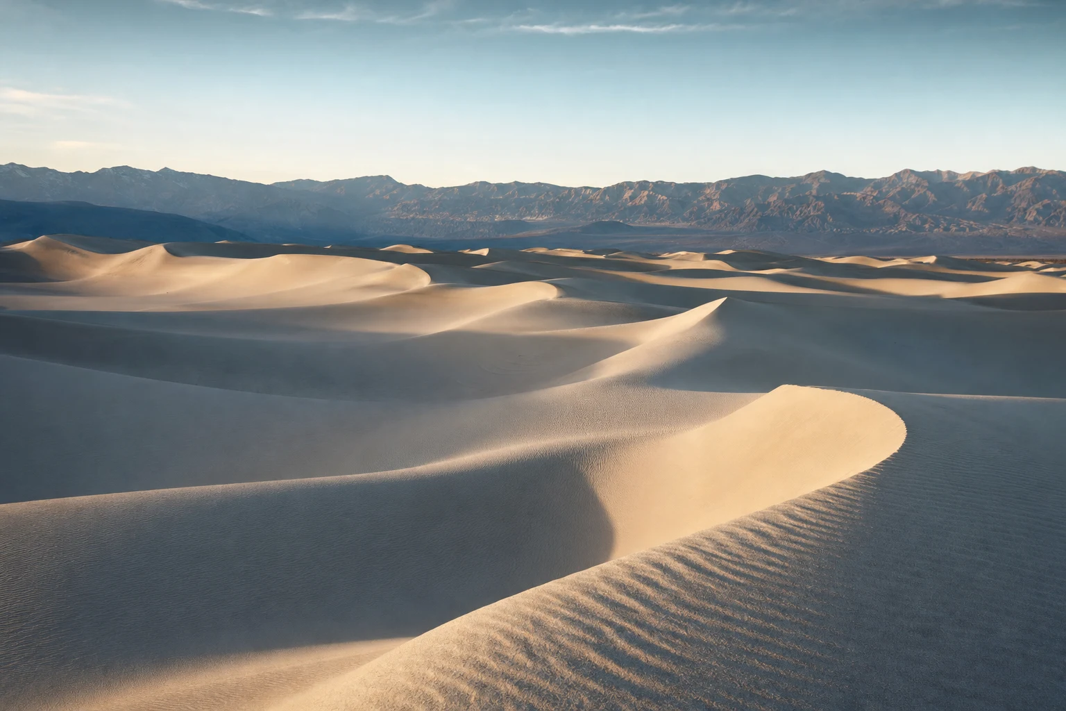 Dunas de arena color marfil con sombras largas de la tarde y un cielo azul pálido